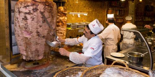 A restaurant worker slices roasted meat from a spit on Istiklal Street, a vibrant, crowded pedestrian street in Istanbul Turkey.